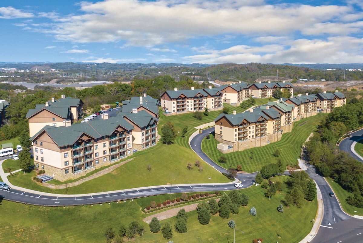 An aerial view captures the expansive layout of the resort, featuring multiple buildings with stone and wood exteriors. Lush green lawns surround the structures, with winding pathways leading through the landscape. Rolling hills and a clear sky define the backdrop.