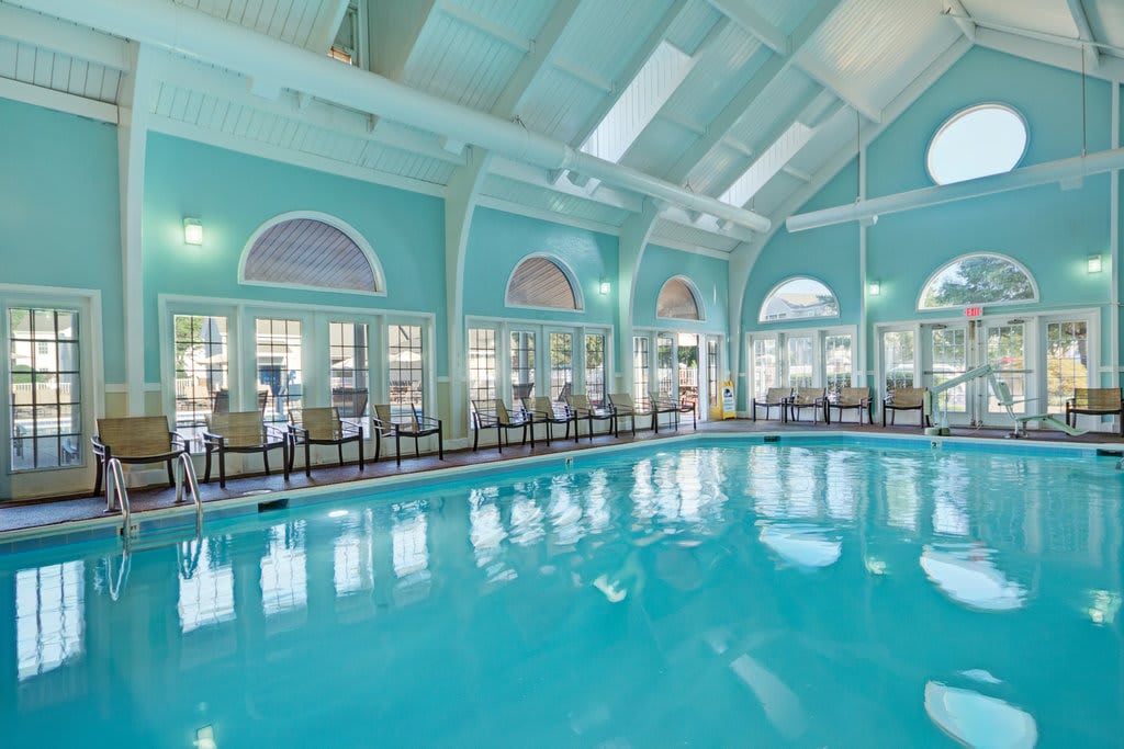An indoor swimming pool is framed by large arched windows allowing natural light to fill the space. The room features a high ceiling and light blue walls, with various seating options available around the pool's edge, creating a serene setting.