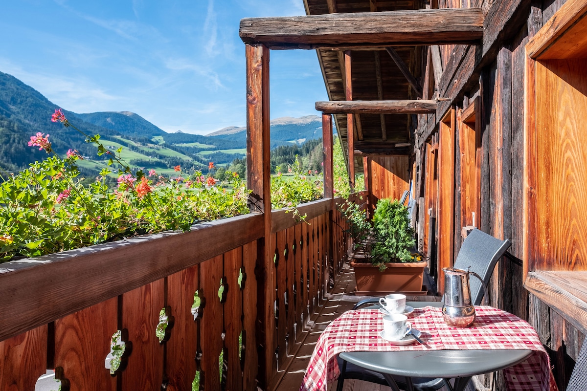 A balcony is featured with wooden railings and decorative cutouts. A table with a checkered tablecloth is set for two, including a coffee pot and cup. Surrounding greenery and mountain views create a serene backdrop, while flowering plants enhance the outdoor space.