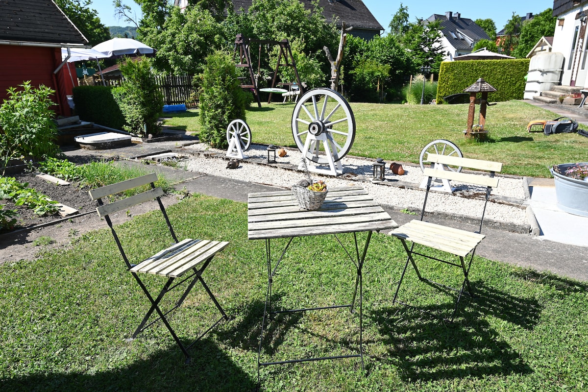 An outdoor seating area features a small table accompanied by two folding chairs, situated on a grassy lawn. Decorative elements, including a wagon wheel and various garden ornaments, are visible in the background, with trees lining the space.