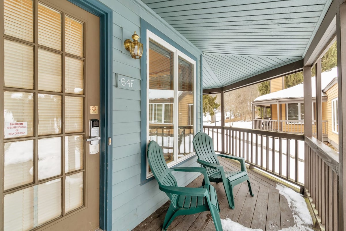 A covered porch is showcased, featuring two green Adirondack chairs positioned on a circular wooden deck. Snow is visible on the ground, with additional buildings in the background reflecting a tranquil resort setting.