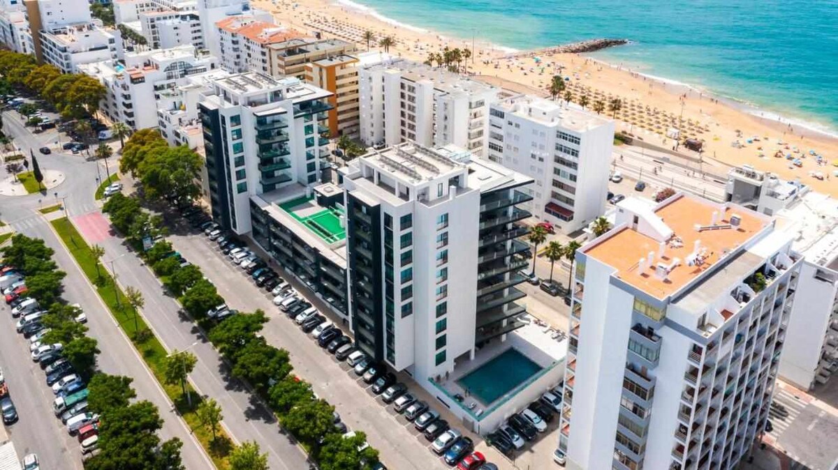 An aerial view captures the modern Terraços de Quarteira building, prominently showcasing its private pool. Texture-rich sandy beaches and the crystalline ocean are visible nearby, along with palm-lined streets and a row of parked vehicles, creating a vibrant coastal atmosphere.