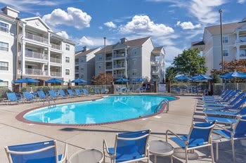 The outdoor swimming pool is surrounded by blue lounge chairs, offering a space for relaxation. Umbrellas provide shade, and a backdrop of light-colored buildings adds to the resort's atmosphere. The sky is bright and partly cloudy, enhancing the inviting setting.