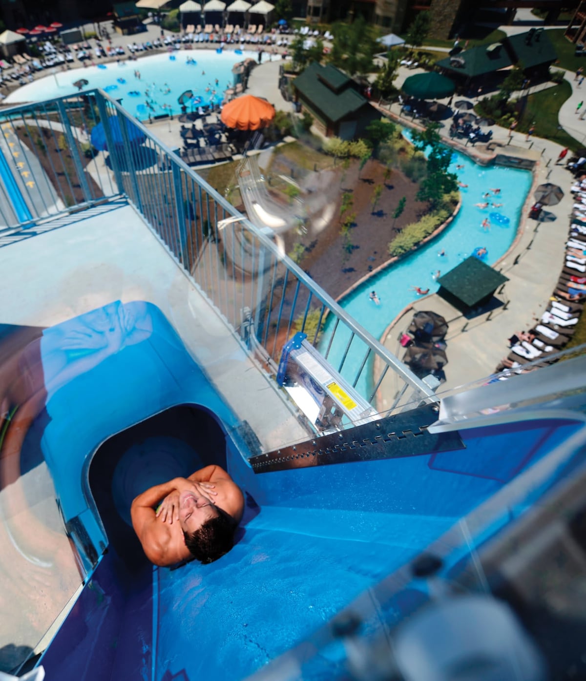A water slide spirals down from a height, leading into a large outdoor water park. Below, guests enjoy a turquoise pool surrounded by sun loungers and shaded seating areas, with vibrant umbrellas providing shade in various colors.
