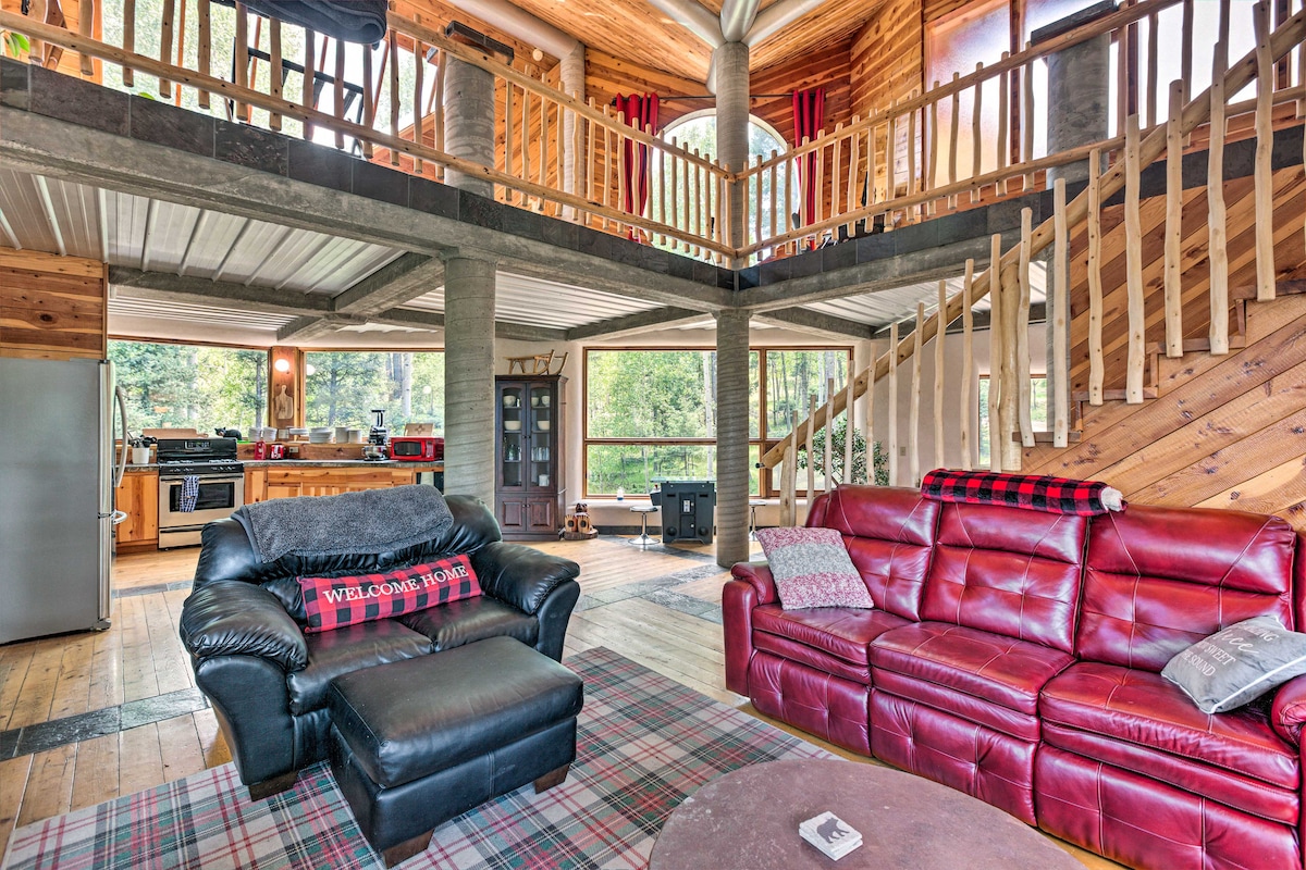 A spacious living area is prominently featured, showcasing a mix of leather seating and a plaid area rug. A staircase leads to a loft area above, while large windows allow natural light to illuminate the room. The open layout connects the living space to the kitchen in the background.
