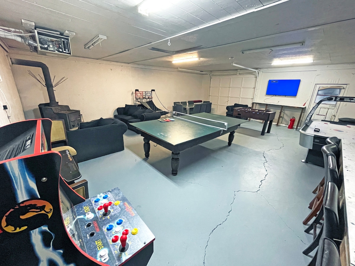 A spacious entertainment room is equipped for leisure activities. A large table stands at the center for games, surrounded by comfortable couches and seating. A classic arcade game console is positioned to one side, while a television screen is mounted on an adjacent wall.