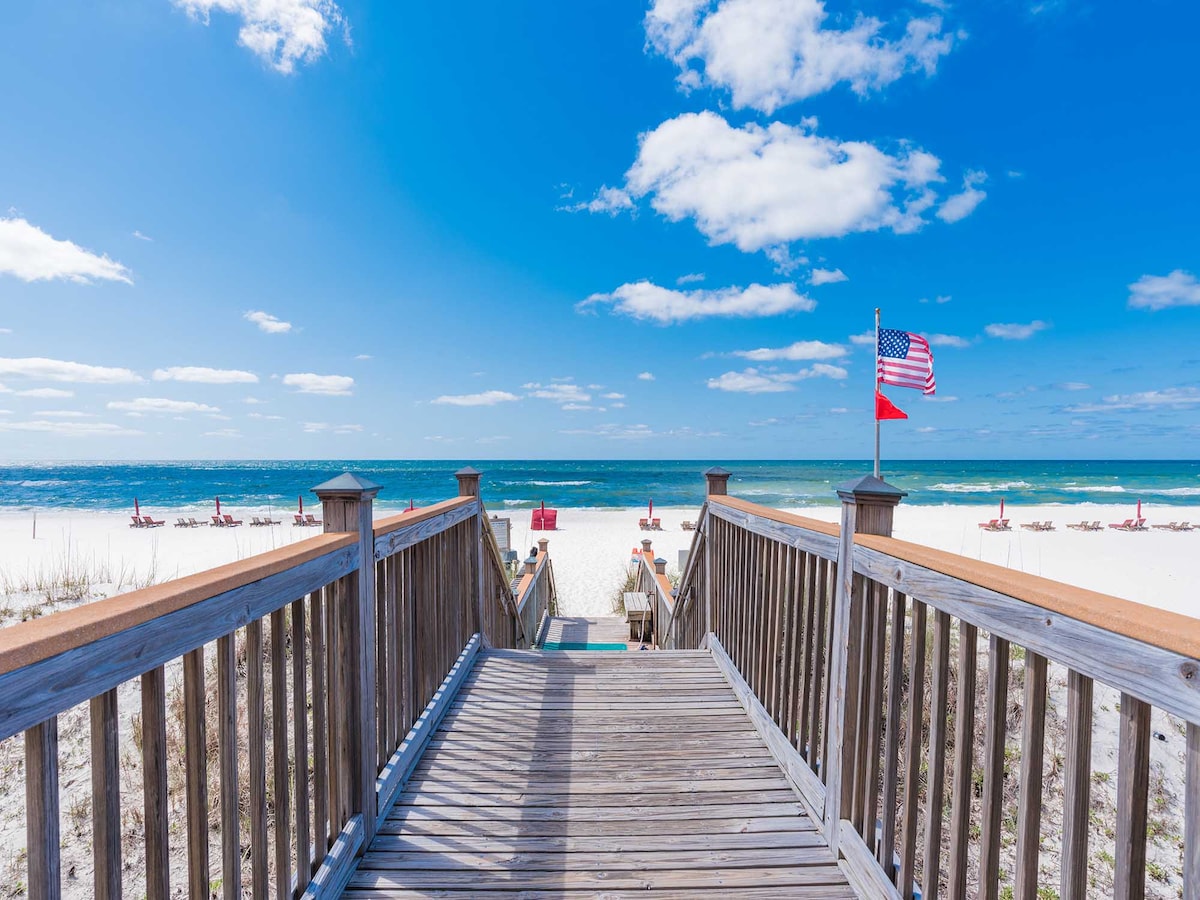 A wooden boardwalk leads to the beach, surrounded by soft sand and rolling waves. The bright blue sky is dotted with fluffy white clouds, while an American flag and a red flag flutter gently in the breeze. Beach chairs are visible in the distance.