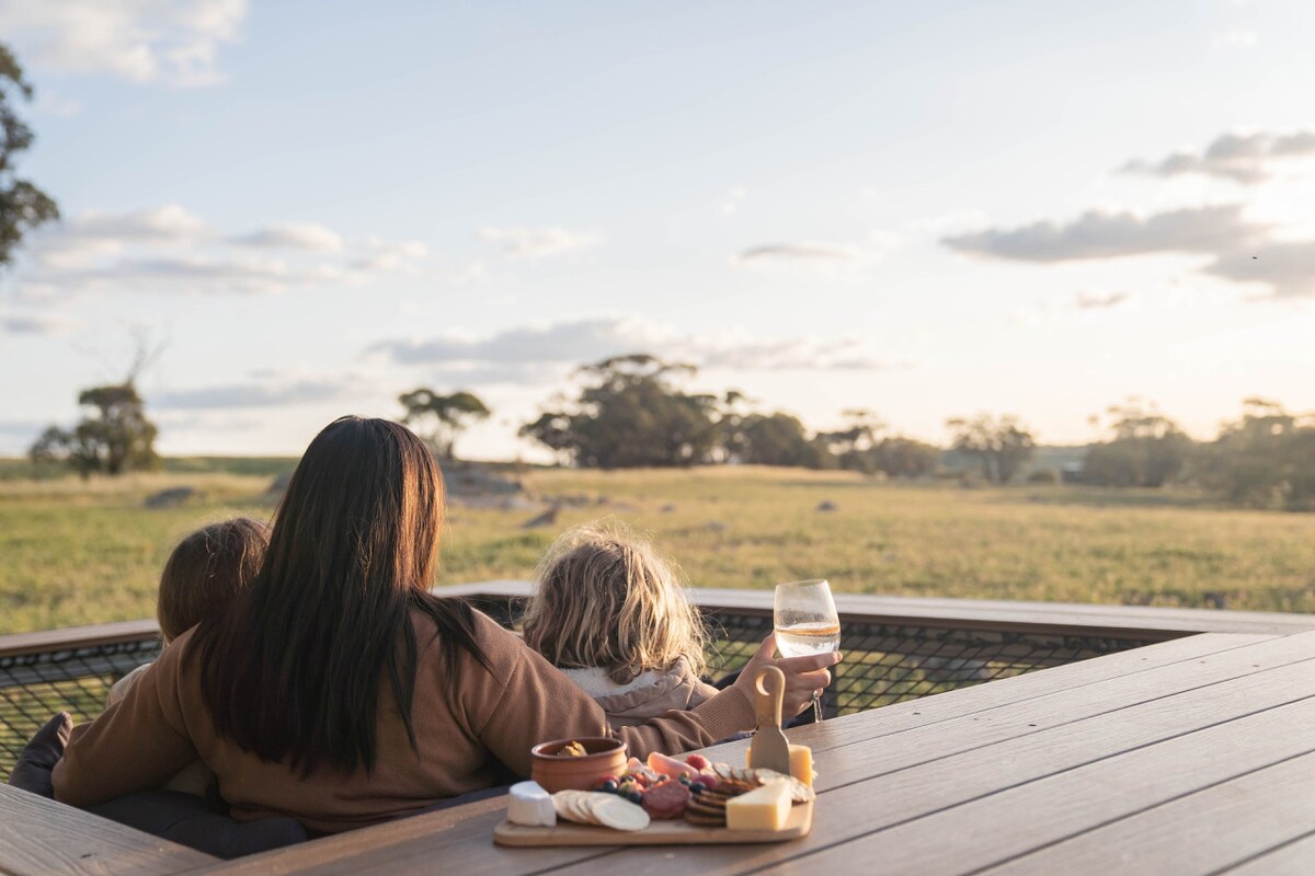 A woman sits on a wooden deck, facing a tranquil landscape of rolling fields. Two children are nestled close. A grazing sheep can be spotted in the distance. An assortment of snacks and a glass of wine are placed on the table nearby, offering a picturesque setting for relaxation.