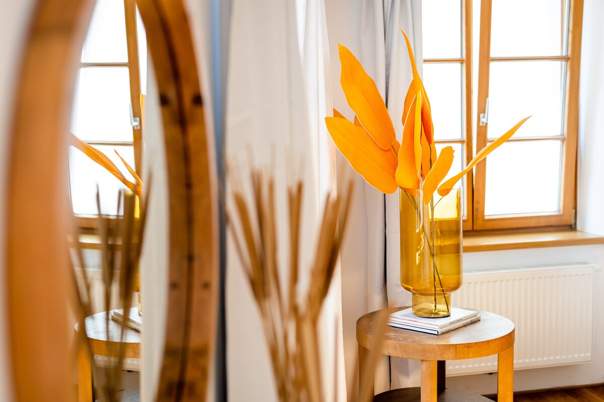 A vibrant arrangement of orange leaves is displayed in a tall glass vase on a wooden side table. The reflection in a large round mirror captures both the floral display and the light streaming in through a window with multiple panes.