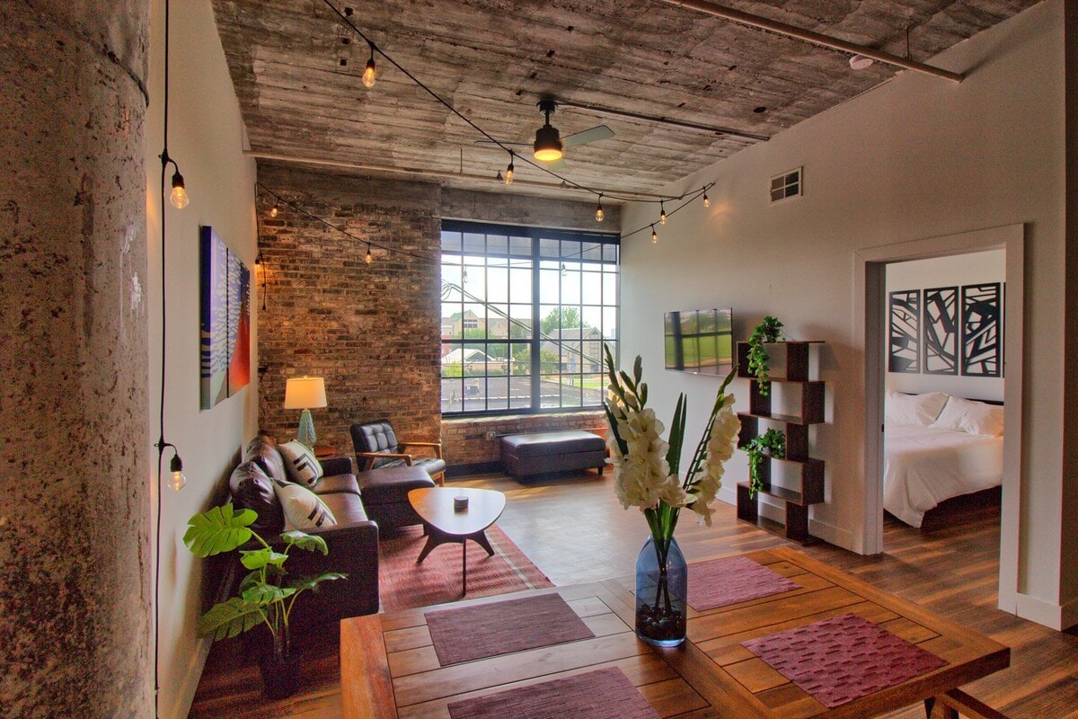 A spacious living area features a combination of wood and industrial elements. A large window with industrial steel framing allows natural light to fill the room. Comfortable seating is arranged around a central coffee table, enhancing the open layout design.