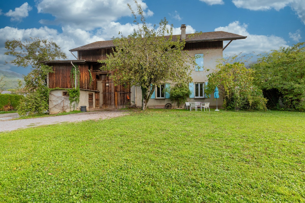 A detached house is displayed, featuring a blend of natural and rustic elements. Lush greenery surrounds the property, while a neatly maintained garden area includes a small table and chairs. Large windows allow for ample natural light to enter the home, enhancing the outdoor view.