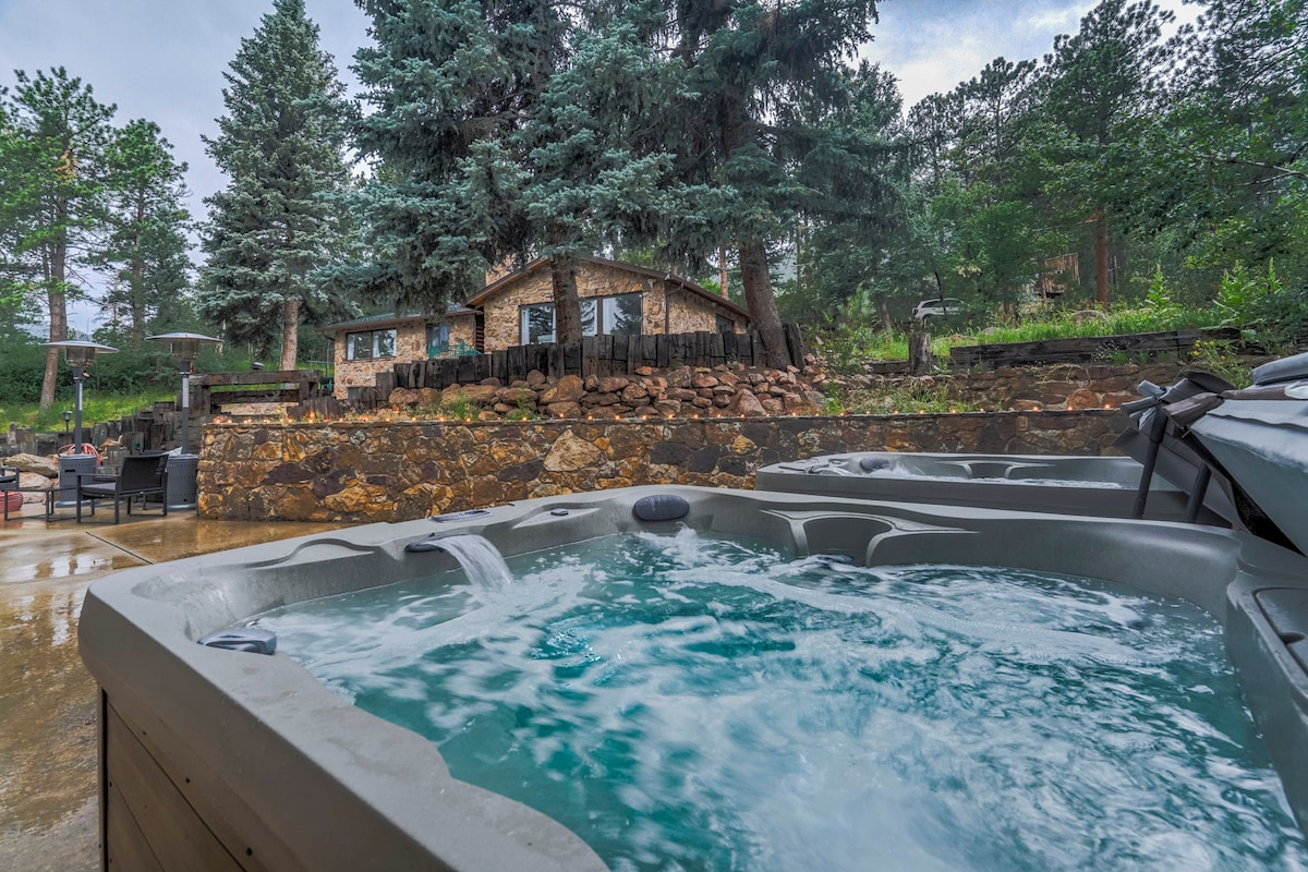 Two shared hot tubs are situated on a stone patio, surrounded by lush trees and greenery. The house stands in the background, showcasing a rustic stone exterior. Seating areas are visible nearby, inviting relaxation amidst the natural surroundings.