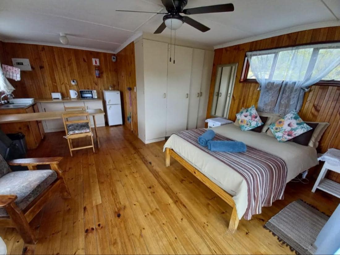 An open plan sleeping area features a spacious bed with layered bedding and decorative throw pillows. Natural wood paneling creates a warm ambiance. A kitchenette with a microwave and refrigerator is visible, alongside a small dining area. A comfortable chair and cabinetry are also present.