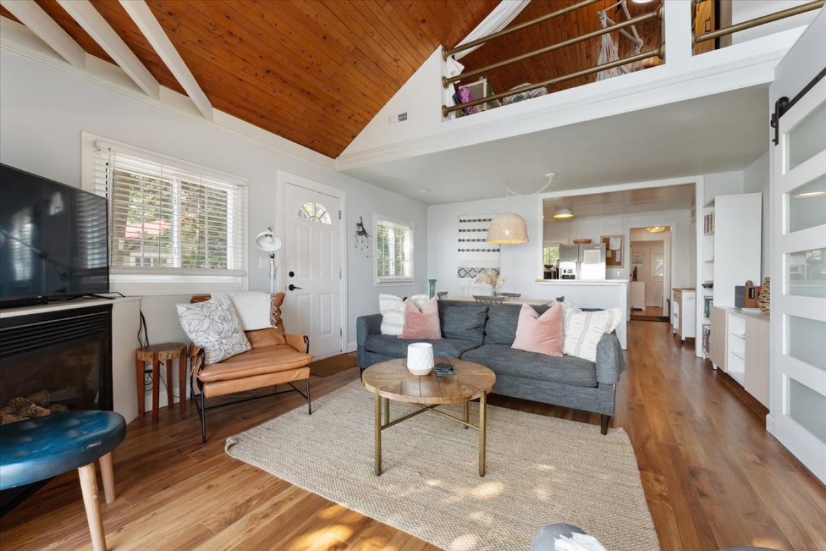 The living area features a comfortable grey sofa adorned with accent pillows, paired with a stylish armchair. A round wooden coffee table sits on a woven rug, while sunlight streams through the large windows, illuminating the space decorated with wooden beams and modern furnishings.