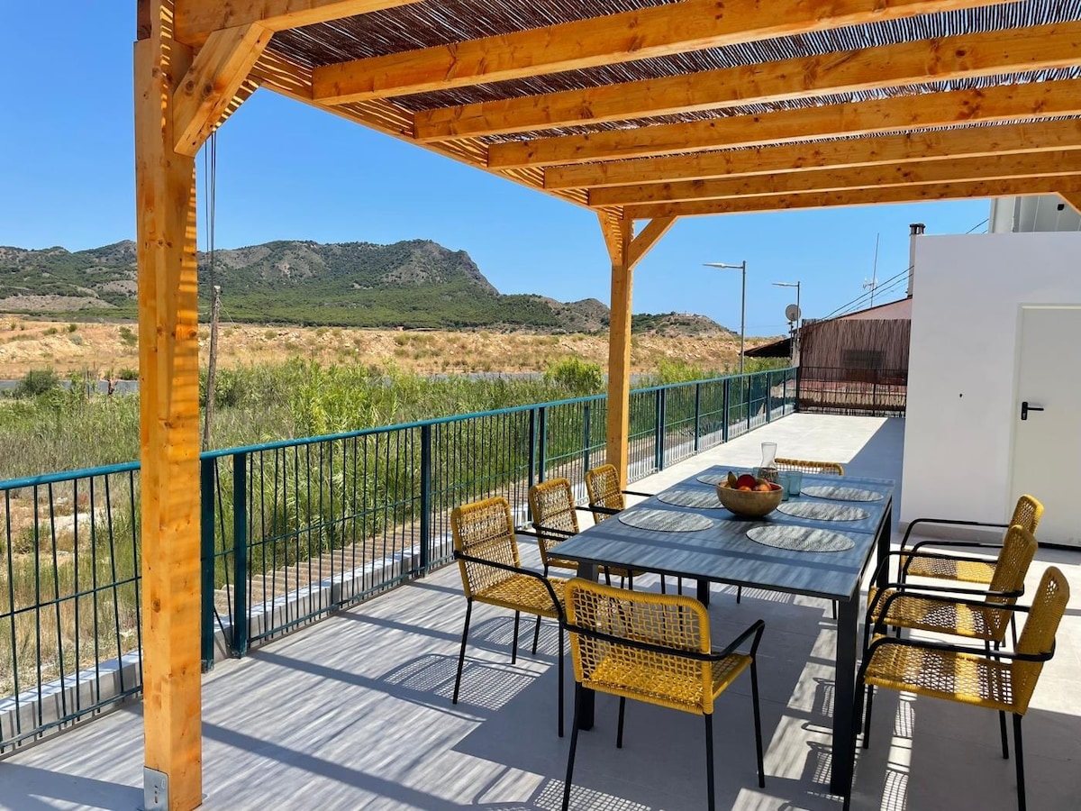 A spacious terrace features a wooden pergola providing shade. A long table surrounded by six chairs is set on a tiled surface. The backdrop includes scenic views of gentle hills and greenery, under a clear blue sky.