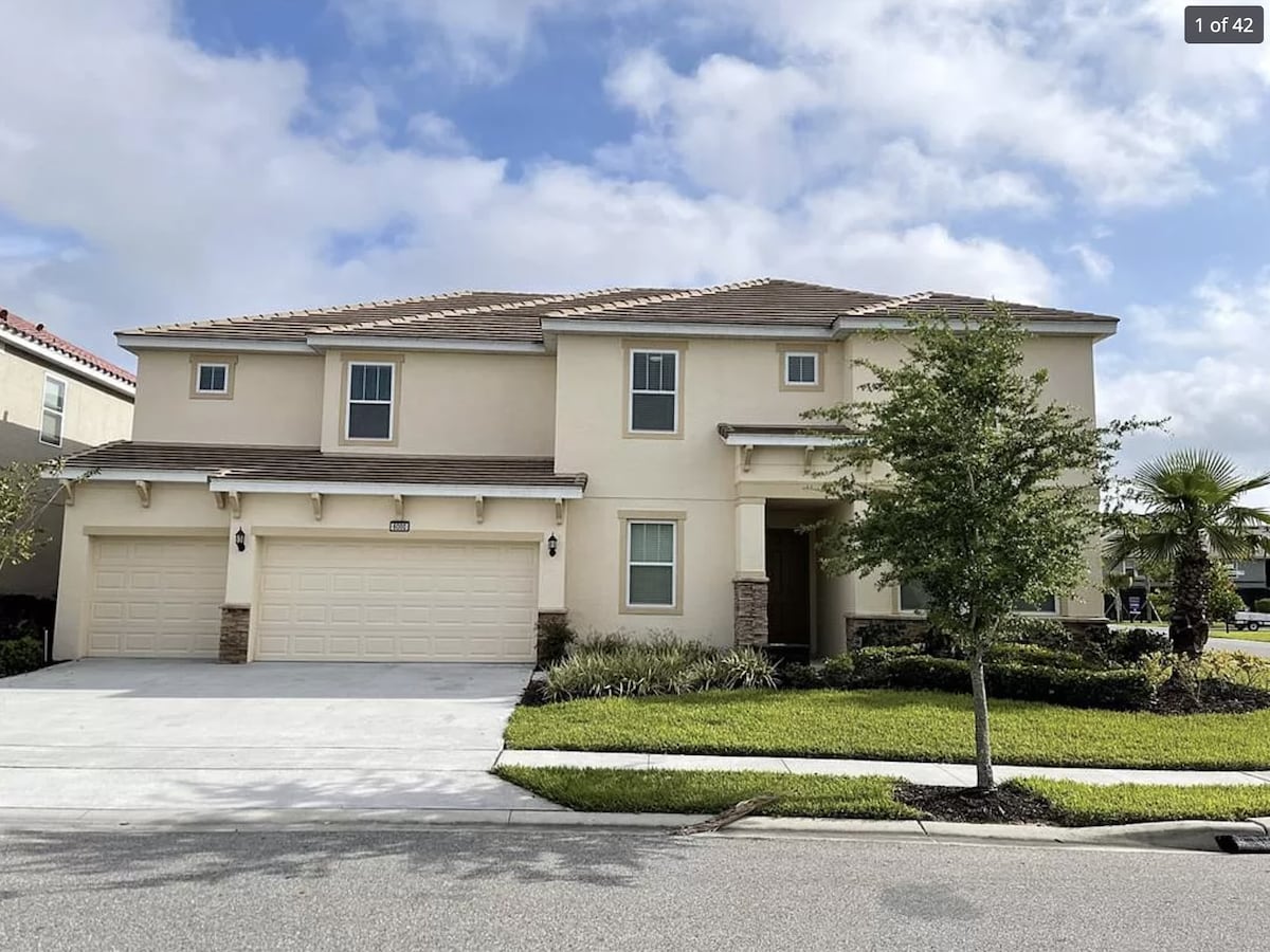 A two-story home is presented with a light-colored exterior, featuring a double garage and welcoming entrance. Lush green landscaping surrounds the property, including shrubs and palm trees. The sky above is partly cloudy, offering a hint of brightness to the scene.