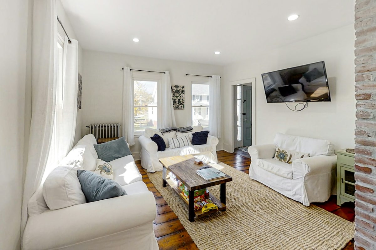 A bright living area is furnished with a plush white sofa set and a wooden coffee table. Natural light floods the space through large windows, while a flat-screen TV is mounted on the wall. A woven rug adds warmth to the hardwood floor.