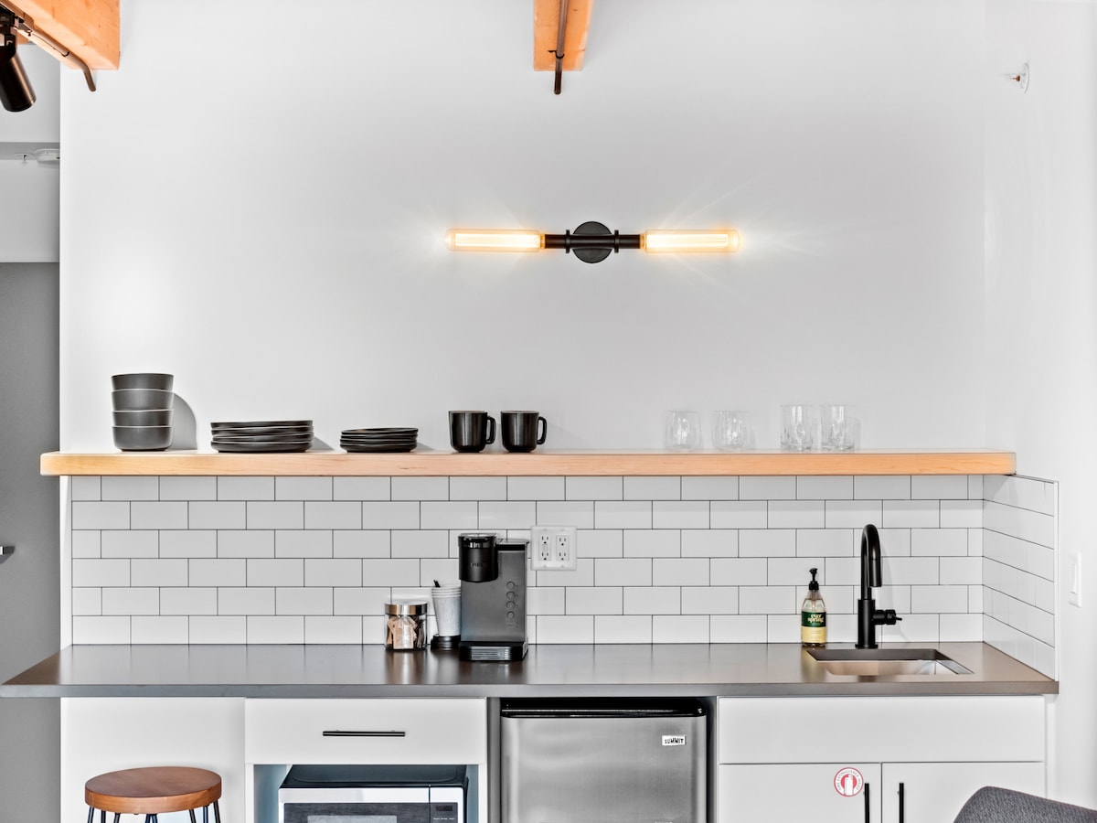 The kitchenette features modern countertops and a polished sink area. Open shelves display dishware, including plates and cups, while a coffee maker and mini fridge occupy the accessible space below. Natural light highlights the contemporary design and warm wood accents on the ceiling.