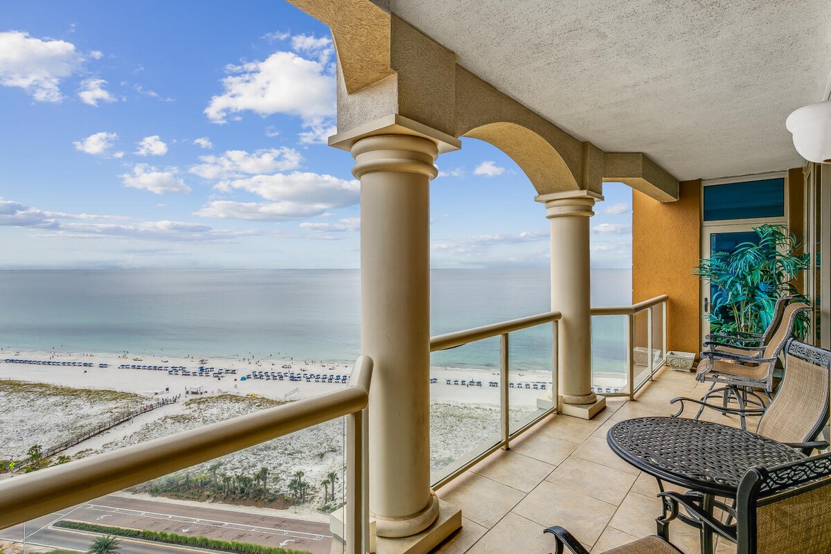 A spacious balcony is featured, offering sweeping views of the Gulf of Mexico and the sandy beach below. Comfortable outdoor seating is arranged, with decorative columns adding an elegant touch. The clear blue sky complements the tranquil water, creating a serene setting.
