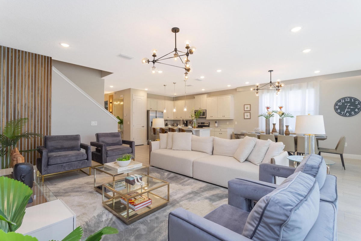 A spacious living area features a modern sectional sofa and two accent chairs, arranged around a glass coffee table. A well-lit kitchen and dining area are visible in the background, connected through an open layout. Decorative lighting and potted plants add a touch of elegance.