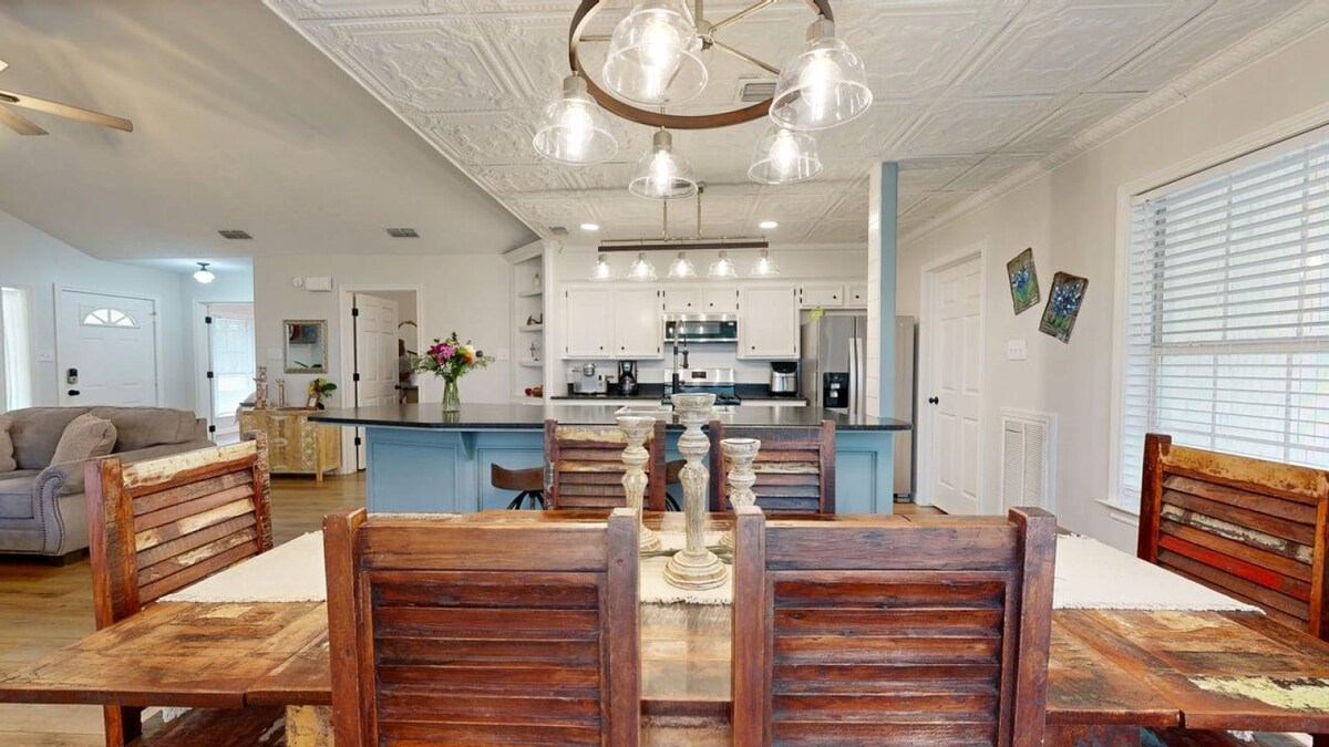 An open concept living and kitchen area features a spacious dining table made of reclaimed wood, flanked by rustic chairs. A large kitchen island is positioned in the background, with modern appliances visible. The ceiling showcases decorative tiles, while natural light enters through multiple windows.
