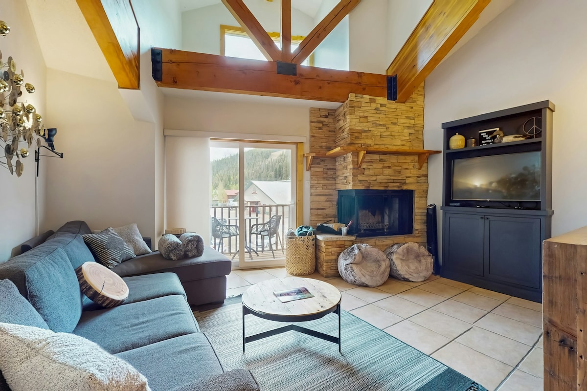 A spacious living area features a modern sectional sofa arranged around a circular coffee table. Natural light fills the room through large glass doors leading to a balcony. A stone fireplace serves as a focal point, complemented by a television on a dark cabinet.