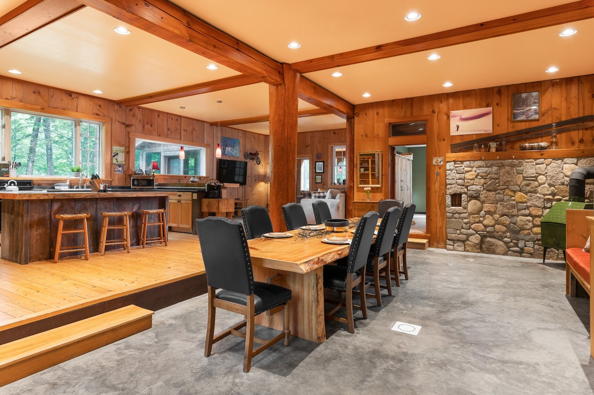 A spacious dining area features a large wooden table surrounded by black chairs, accommodating several guests. Natural light pours in through expansive windows, illuminating the warm wooden walls and flooring. A well-equipped kitchen is visible in the background, with a rustic stone fireplace accentuating the charm.