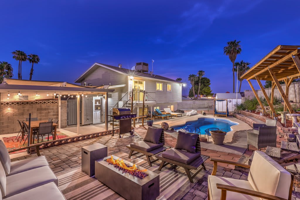 The outdoor area features a well-lit pool surrounded by comfortable lounging chairs and a fire pit. A spacious cabana offers shade, while additional seating is arranged for relaxation. The house, visible in the background, showcases a simple, modern design set against a twilight sky.