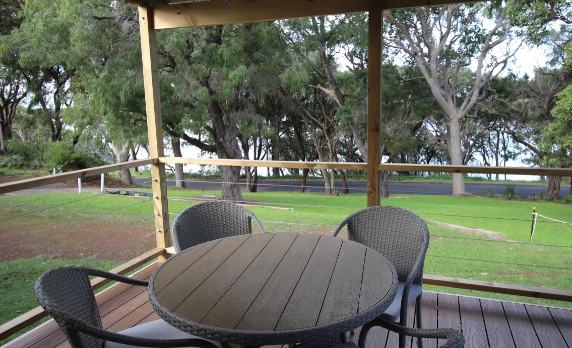 A covered balcony features a round wooden table surrounded by four wicker chairs. The space overlooks a lush green lawn and mature trees, creating a serene outdoor area ideal for relaxation or dining.