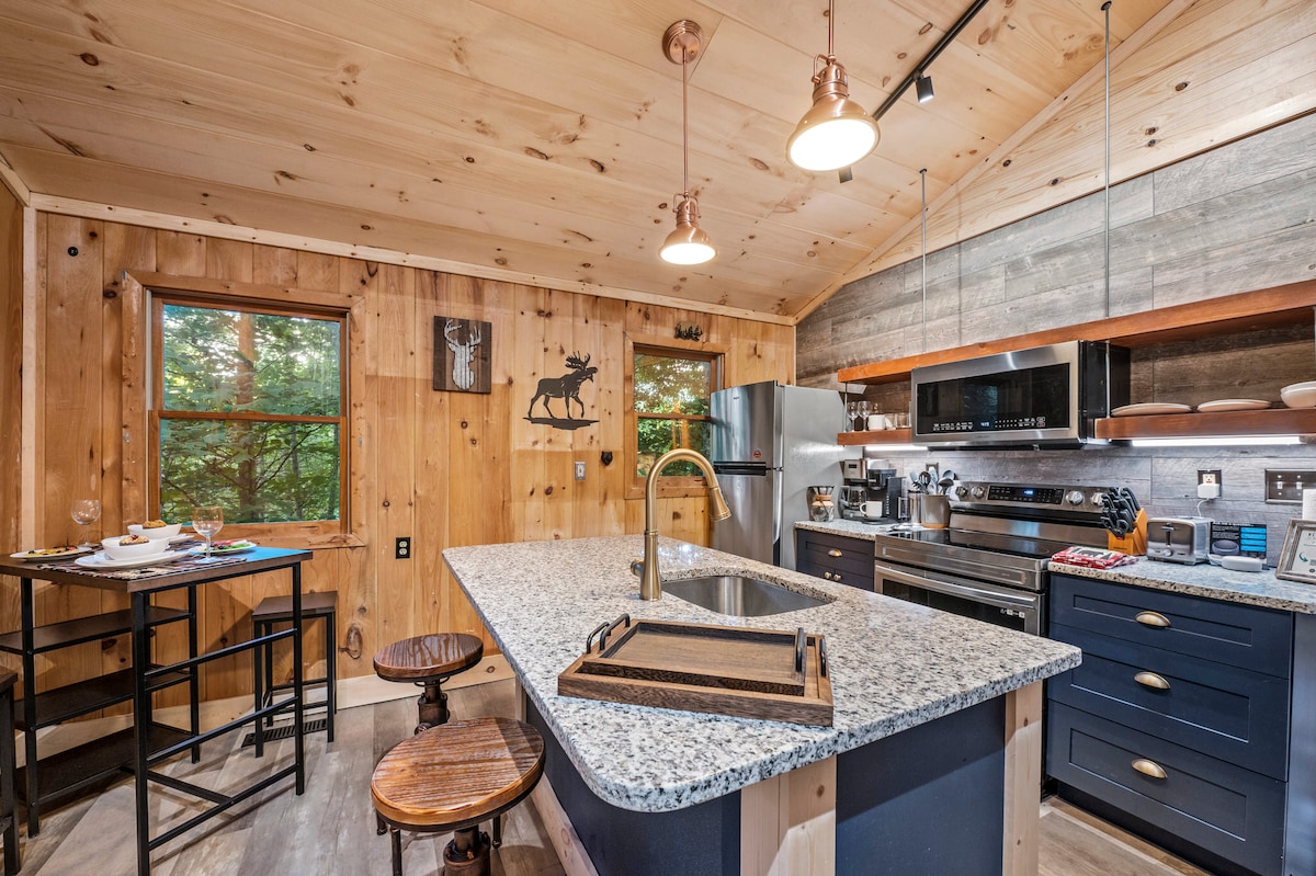 A fully equipped kitchen features granite countertops and modern appliances, providing a functional space for meal preparation. A high-top table and stools are positioned nearby for casual dining. Natural light filters through the window, highlighting the warm wood paneling throughout the cabin.