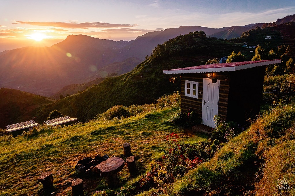 A small cabin with a white door is positioned on a grassy slope, surrounded by lush greenery. The sun rises behind distant mountains, illuminating the landscape. A firepit with seating made from logs sits nearby, adding to the serene outdoor setting.