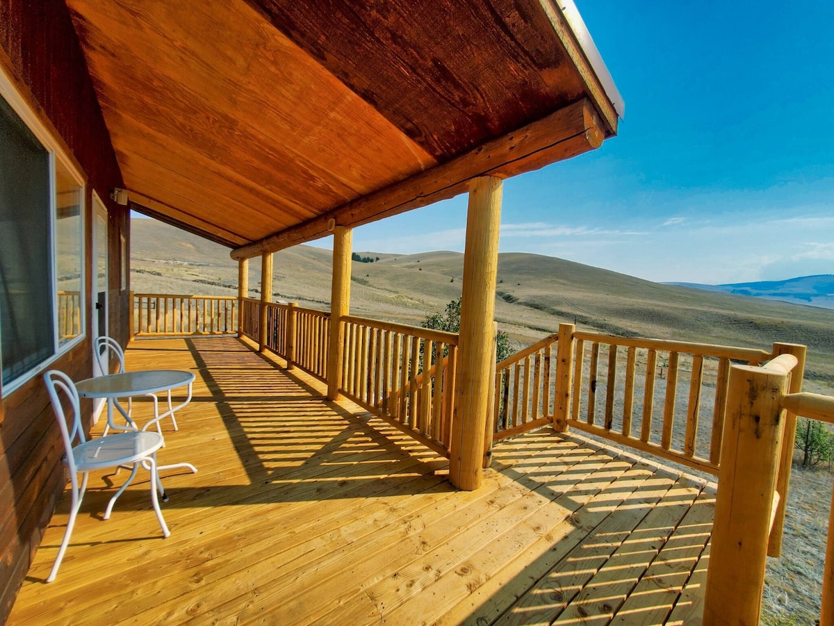 A spacious wraparound deck features wooden flooring and log railing, providing an open view of rolling hills under a clear blue sky. Two white chairs and a small round table are positioned on the deck, inviting guests to enjoy the outdoor scenery.