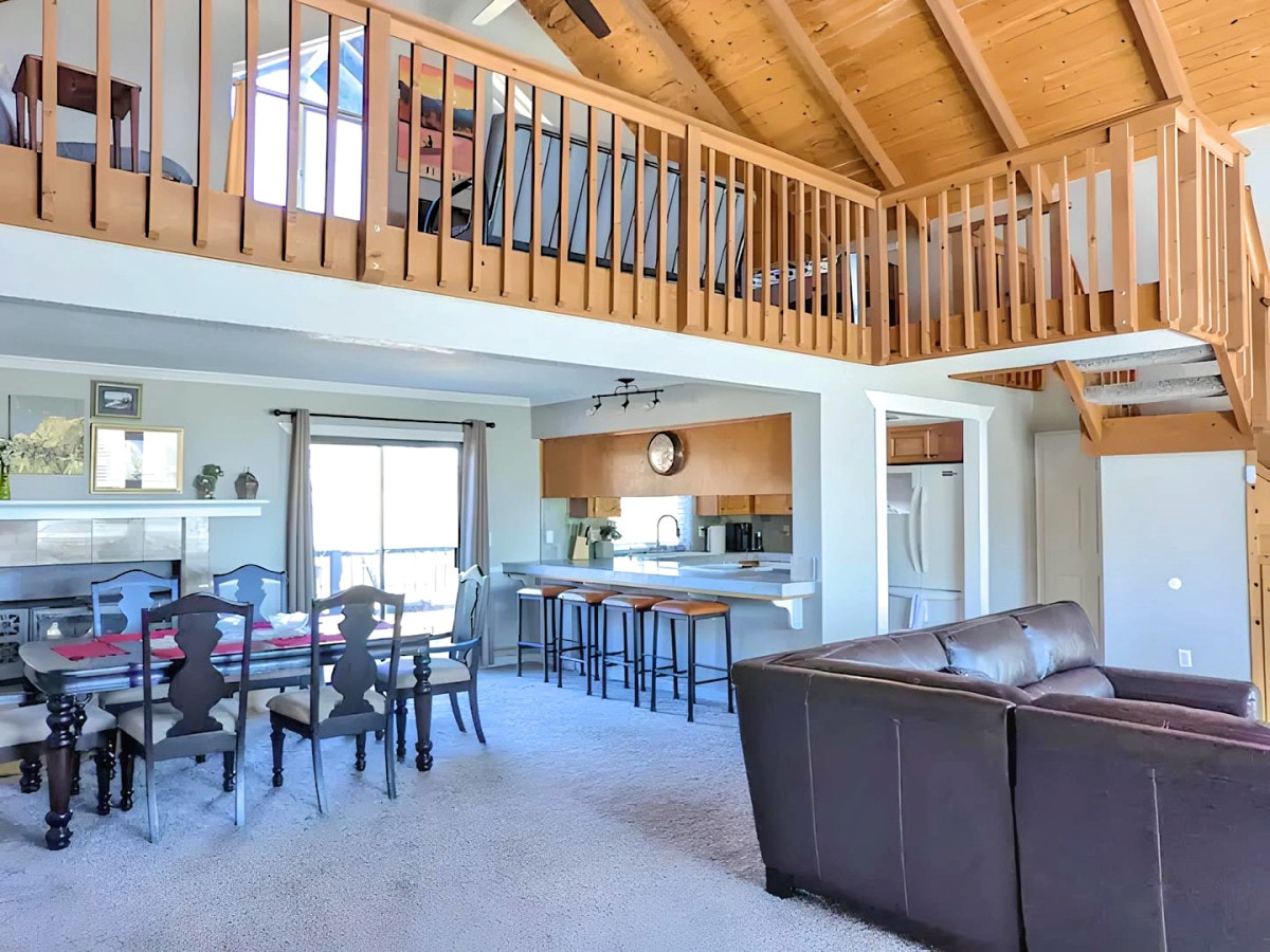 An open floor plan is showcased, featuring a spacious living area with a dark leather sofa and a dining table with seating. A modern kitchen is visible in the background, complemented by high ceilings and wooden beams. A loft area is accessible via a staircase.