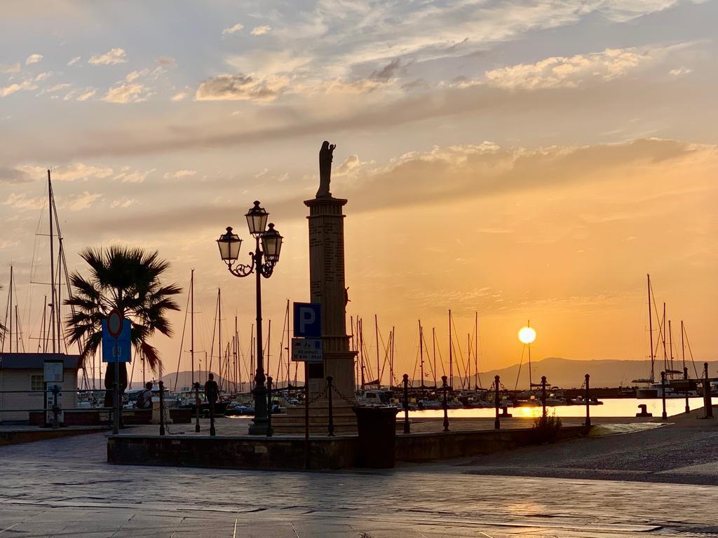 A serene sunset is depicted over a marina, with sailboats anchored in the background. A statue stands prominently, flanked by a palm tree. Warm hues of orange and yellow in the sky contrast with the silhouettes of the boats, creating a tranquil atmosphere.