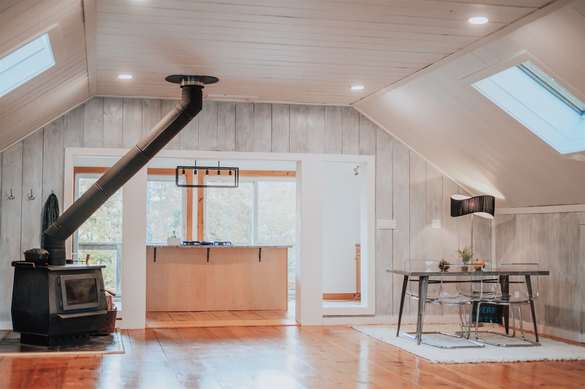 An open-concept living area features a modern wood-burning stove and large skylights, allowing natural light to fill the space. A dining table sits in the foreground, with a minimalist kitchen area visible through the glass doors, enhancing the airy feel of the chalet.