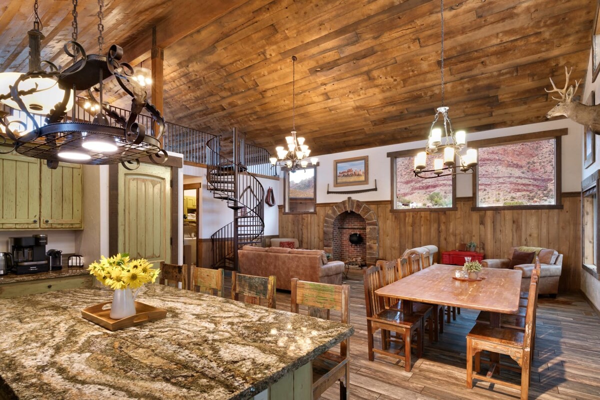 An open-concept area features a large dining table surrounded by rustic chairs, with a stone fireplace at its center. A modern kitchen is visible, showcasing a dark granite countertop and wooden cabinetry. Natural light brightens the space through multiple windows, enhancing the warm wood ceiling.