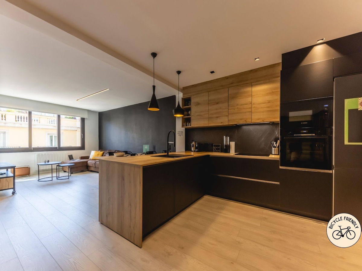 A modern kitchen space features sleek black cabinetry with wooden accents. A kitchen island with a sink and seating area is positioned in the center, contributing to an open layout. Large windows provide natural light, illuminating the adjacent living area furnished with a comfortable sofa.