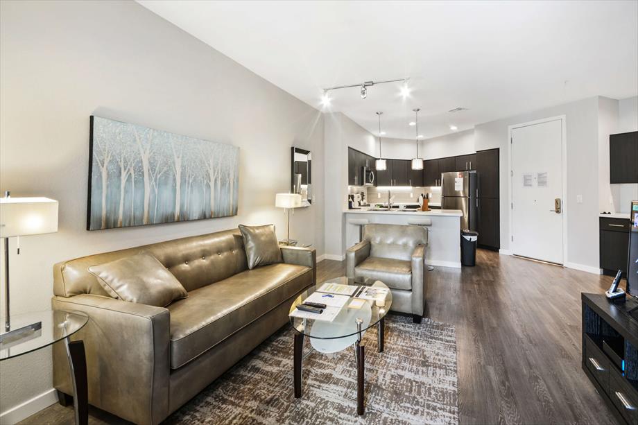 A modern living area features a plush metallic gold sofa paired with a glass coffee table on a patterned rug. The open kitchen displays sleek cabinetry and stainless steel appliances, while soft lighting enhances the neutral tones of the space.