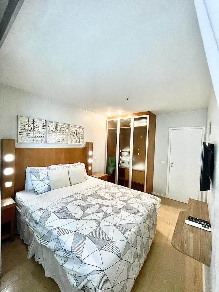 A bright bedroom features a spacious bed with a patterned comforter, positioned against a wooden headboard. On either side, wall-mounted lights provide soft illumination. A wooden wardrobe stands nearby, and a panelled door leads to an additional room, while a television is mounted on the wall.