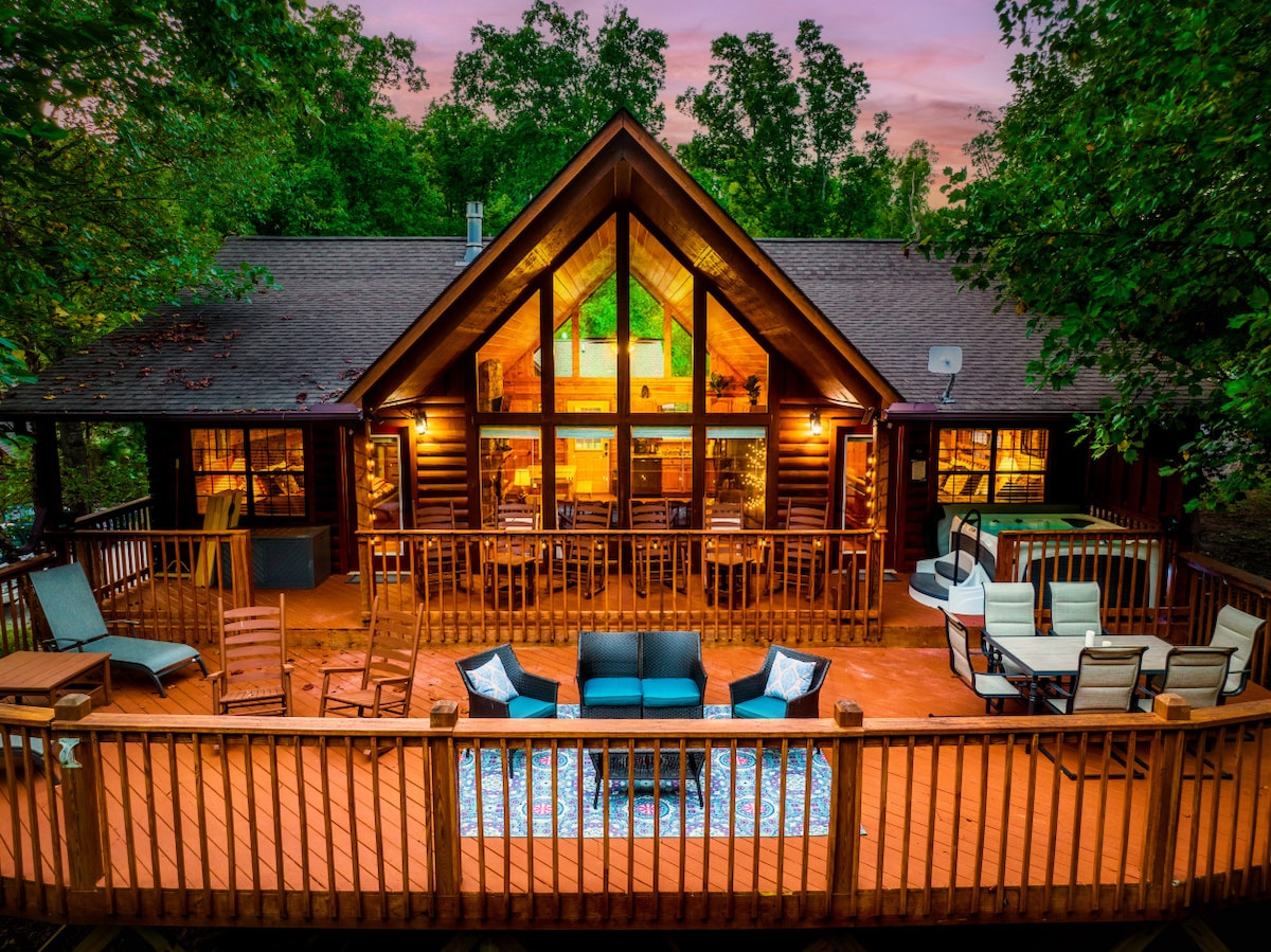The cabin's exterior is framed by lush trees, showcasing a spacious wooden deck with multiple seating arrangements. Large windows reflect the warm glow of interior lights, creating a cozy ambiance as twilight descends. An inviting area rug adds color to the outdoor space.