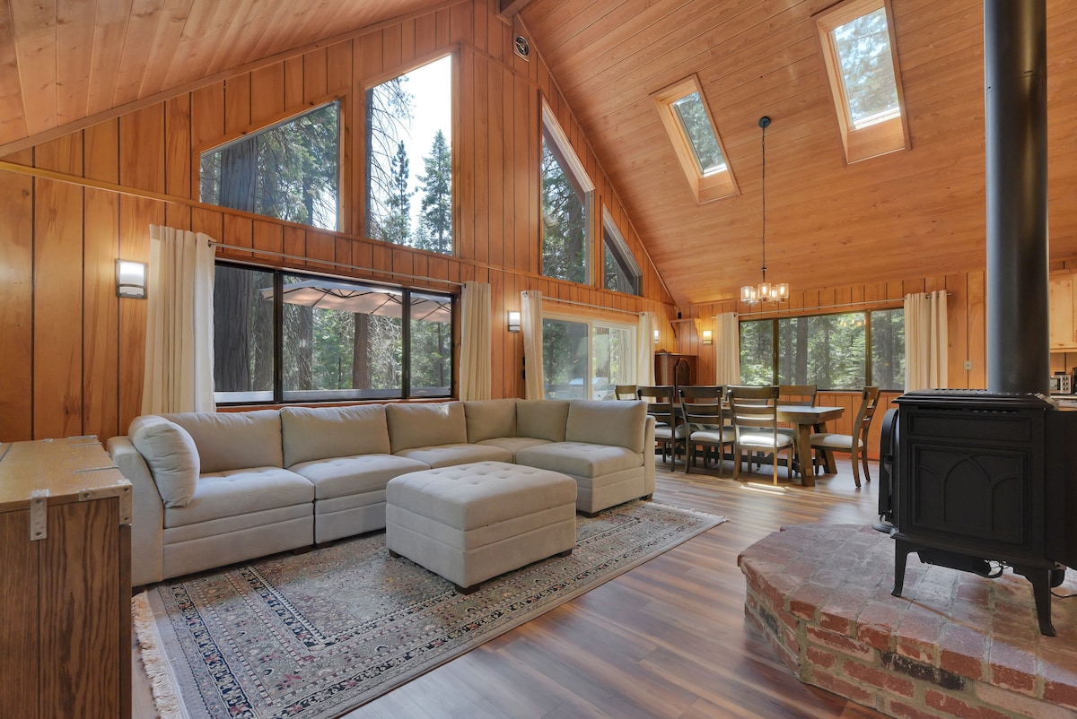 A spacious living area features a large sectional sofa with an ottoman, positioned on a decorative area rug. Natural light enters through multiple windows, showcasing wood paneling and a cozy gas log stove. A dining area and updated kitchen are visible in the background.