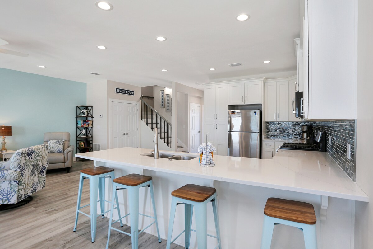 The open kitchen features a large island with seating for four and modern bar stools. Stainless steel appliances and white cabinetry complement the sleek countertop. Light illuminates the space, enhancing the airy atmosphere that flows into the adjoining living area.