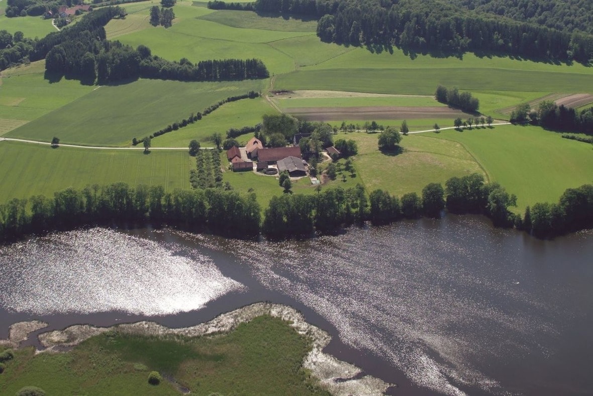 An aerial view captures a spacious landscape, featuring rolling green fields and a gently flowing river. The water reflects sunlight, adding a shimmering effect. A cluster of buildings is situated near the riverbank, surrounded by lush trees and open fields.