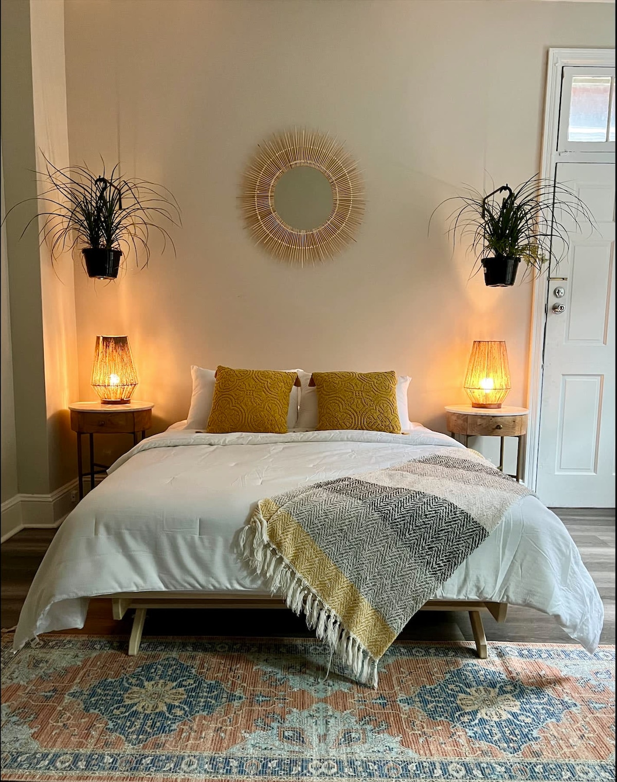 A cozy sleeping area features a spacious bed with white linens and decorative cushions. Two woven lamps cast a warm glow, while potted plants are positioned above the nightstands. A textured throw blanket drapes over the foot of the bed, complemented by an area rug with intricate patterns.