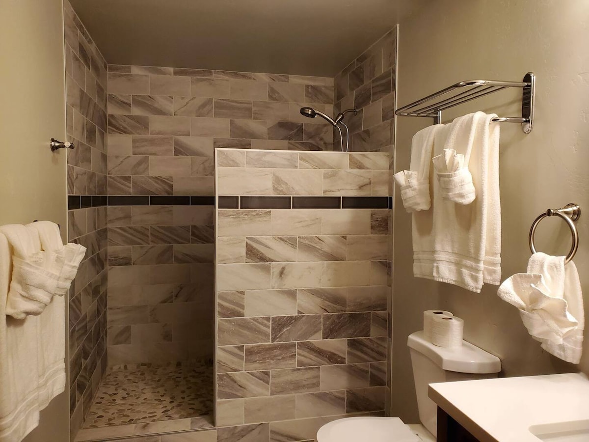 A modern bathroom features a spacious walk-in shower with tiled walls in neutral tones. Soft, white towels hang neatly on racks. A toilet is positioned adjacent to a countertop with a roll of toilet paper, creating a clean and functional space.