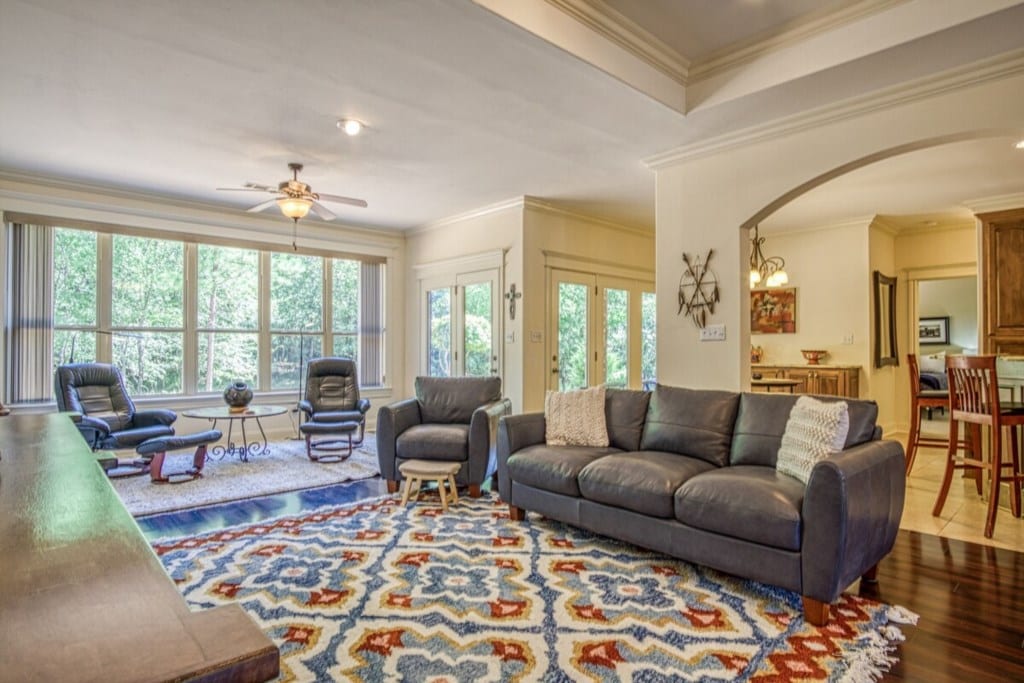 A spacious living area features a large, comfortable sofa with decorative pillows. Two armchairs and reclining chairs provide additional seating. Bright natural light filters through expansive windows overlooking lush greenery. A colorful area rug adds warmth to the hardwood floor, enhancing the inviting atmosphere.