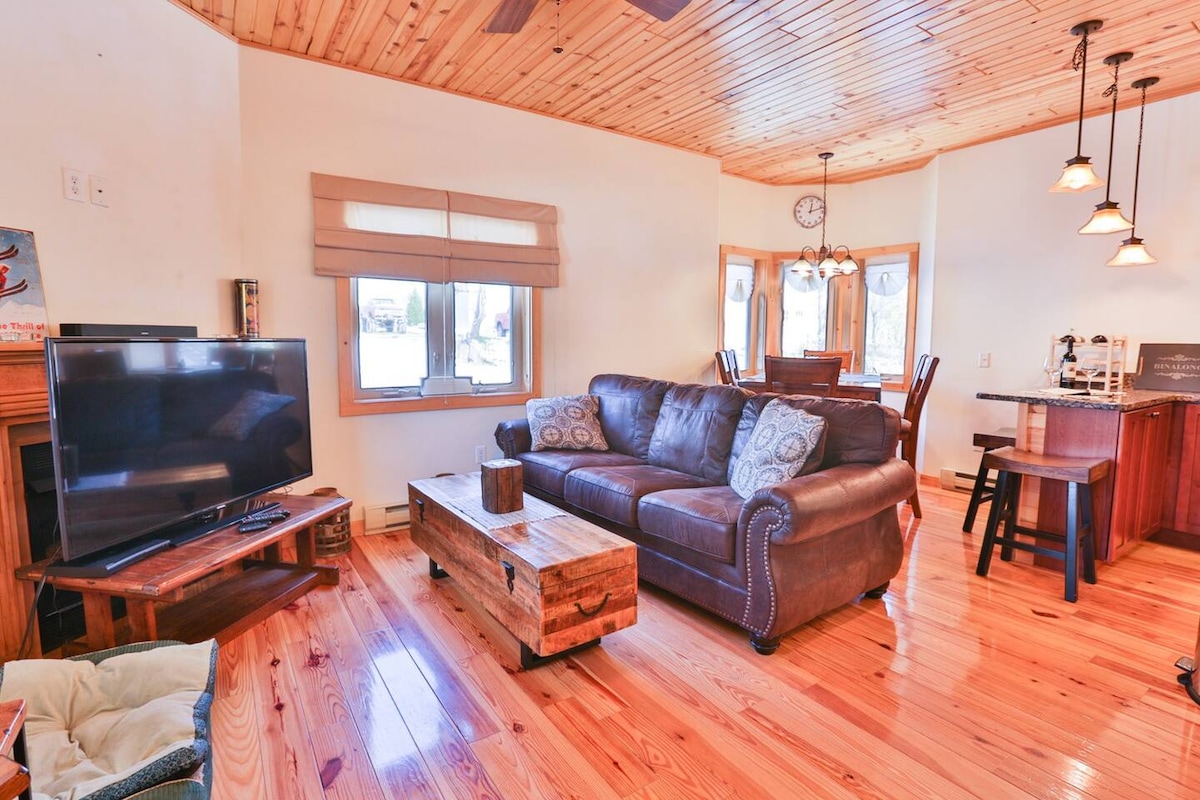 A cozy living area features a leather sofa centered around a wooden coffee table, with a television positioned nearby. Large windows allow natural light to illuminate the space, highlighting the warm wooden ceiling and polished floor. Kitchen bar stools are visible in the background.