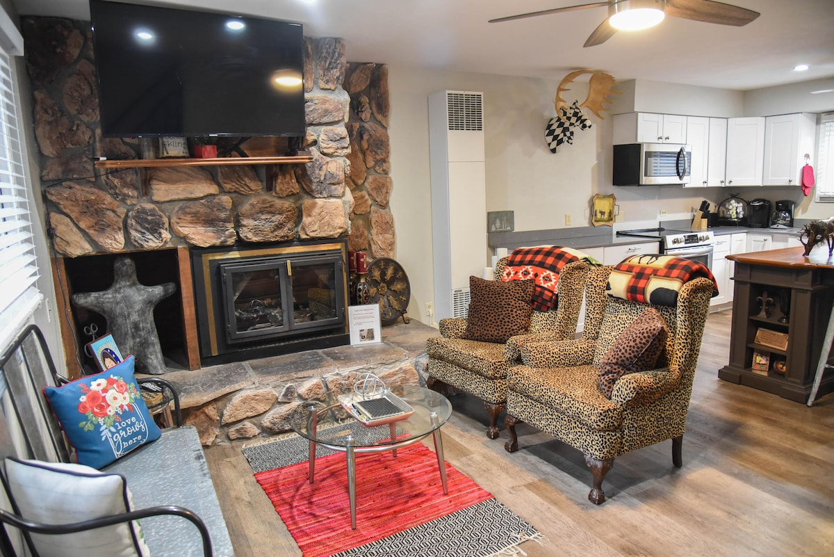 An open living area is presented, featuring a gas fireplace surrounded by stone. Two patterned armchairs and a glass coffee table create a cozy seating arrangement. A flat-screen TV is mounted above the fireplace, and the kitchen is visible in the background with modern appliances.