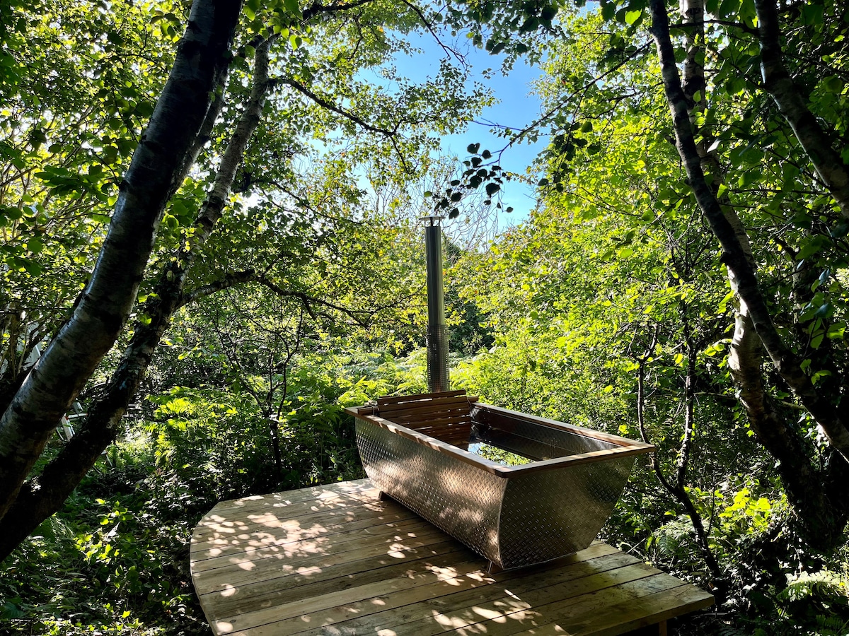 A secluded wood-fired hot tub is positioned on a wooden deck, surrounded by lush greenery and trees. Sunlight filters through the leaves, casting dappled shadows on the deck, creating a serene atmosphere for relaxation in nature.