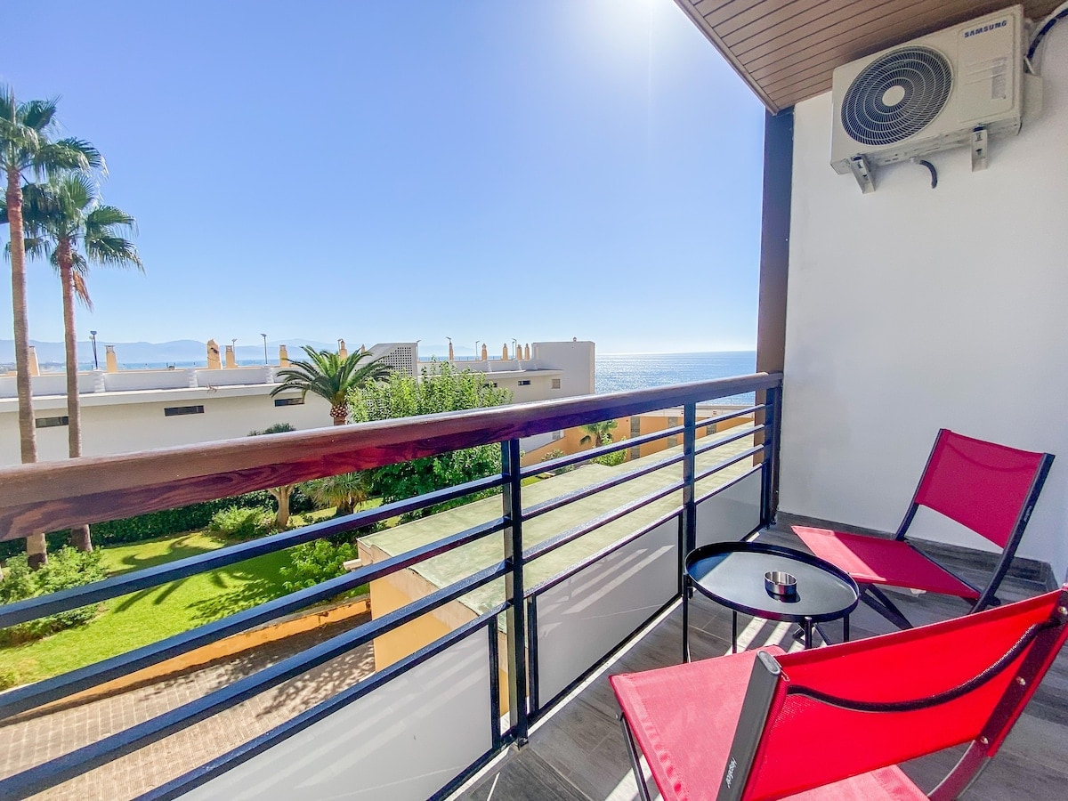 A private terrace is presented with two red chairs and a small round table, overlooking views of the ocean and surrounding greenery. Sunlight bathes the space, creating a bright and inviting environment. An air conditioning unit is mounted on the wall nearby.
